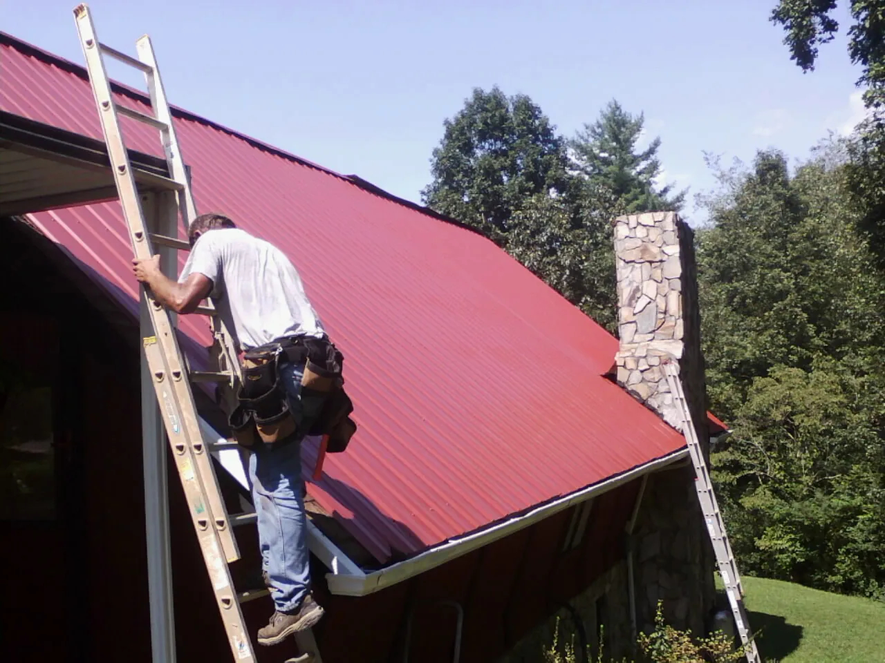 Roofer on a ladder beside a red metal roof and stone chimney — Soffit & Fascia Repair by Roofing Brothers in Ridgefield Park